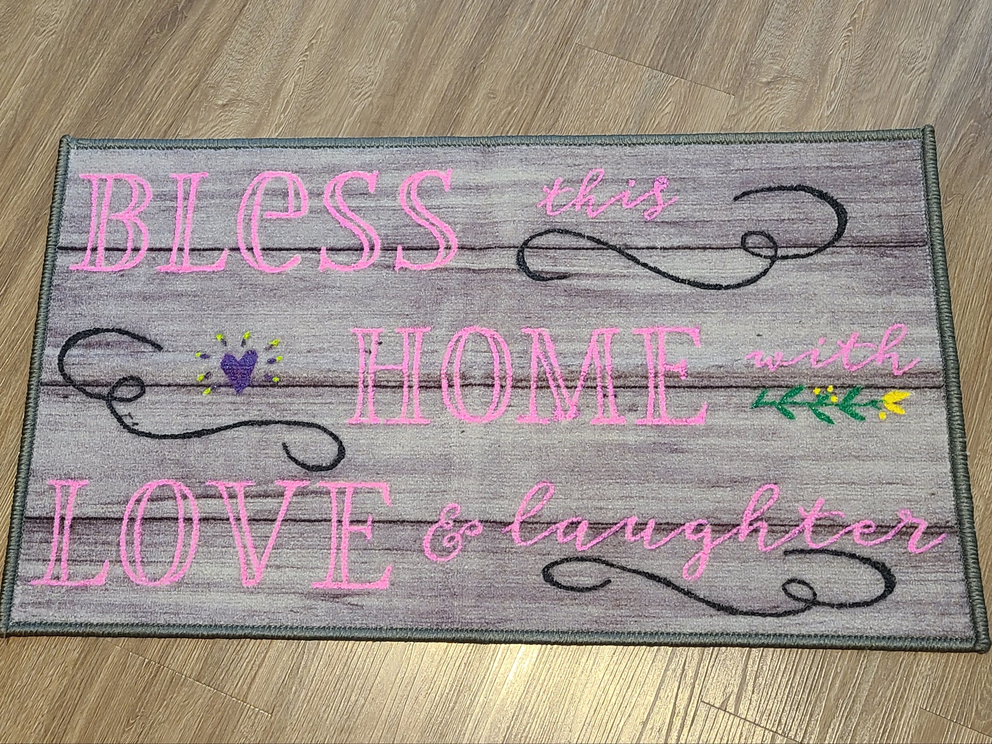 Decorative mat with text 'Bless this home with love & laughter' on a wooden floor.